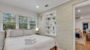 A bedroom with a large bed, white bedding, a brick accent wall, and a built-in shelving unit with plants. - Restful Haven Near Savannah - King Bed (Pooler)