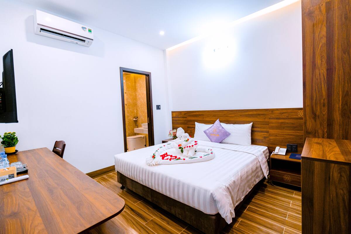 A hotel room with a wooden headboard, a neatly made bed with a floral arrangement, a wooden table, and an air conditioning unit on the wall.