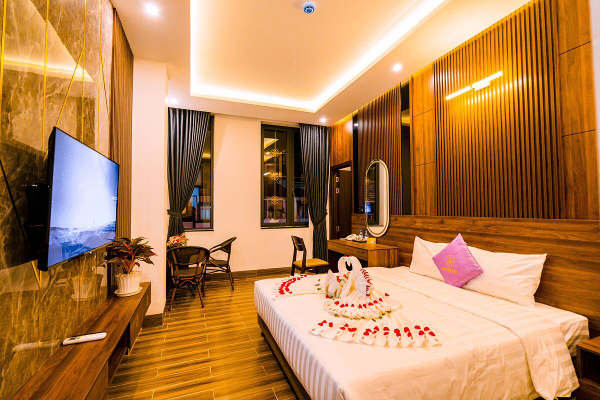 A hotel room with a wooden headboard, a flat-screen TV mounted on the wall, a bed with white bedding and a decorative swan made of flowers, a small table with a plant, and a seating area with chairs.