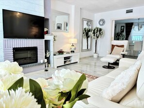 A modern living room with a fireplace, white sofas, a flat-screen TV, and decorative plants.