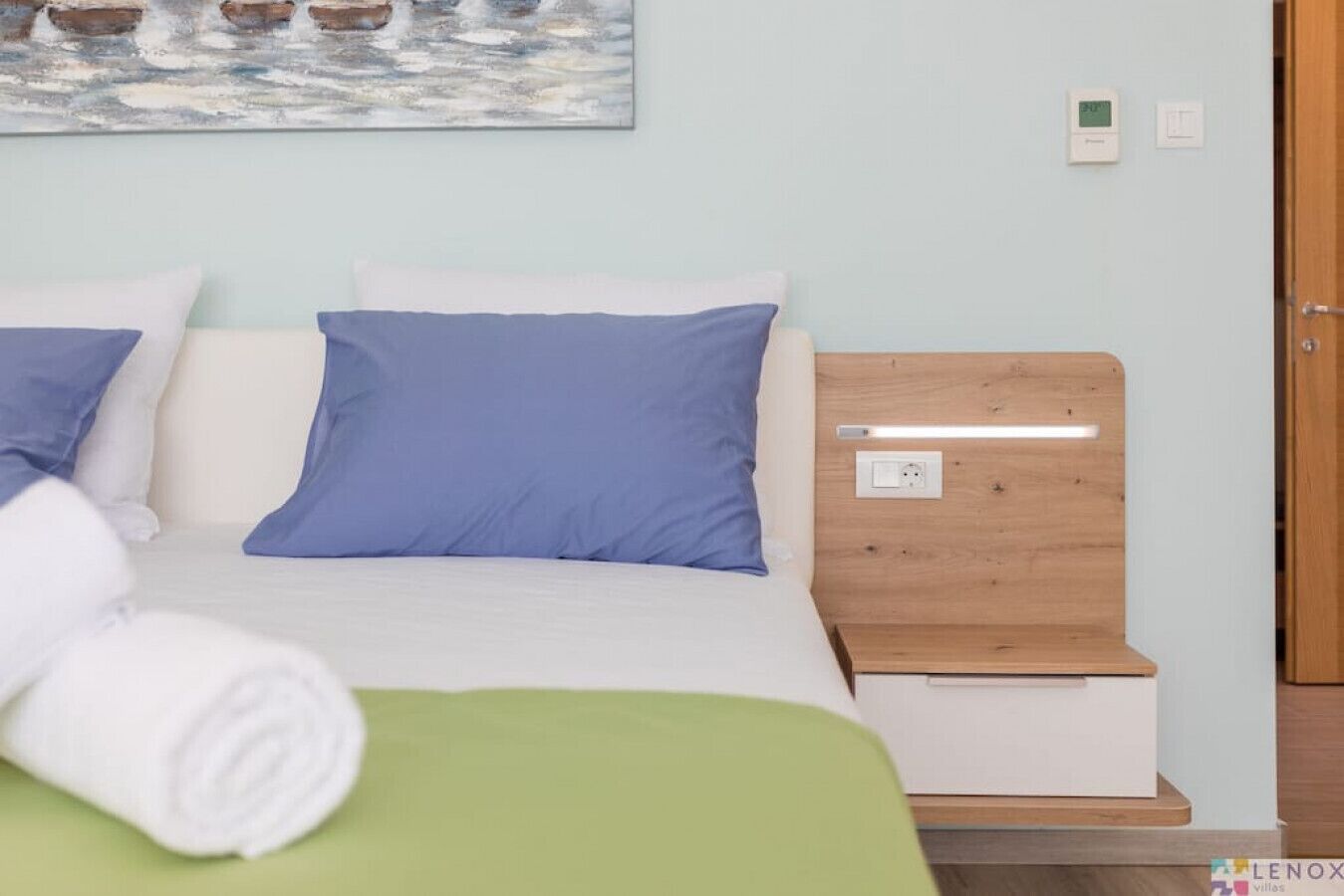 A neatly made bed with blue and white pillows, a green throw blanket, and a wooden nightstand with a lamp.