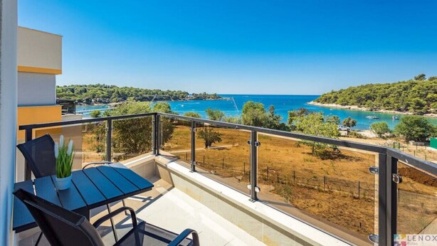 En balkon med bord og stole, der ud til et kystlandskab med klart, blåt vand og grønt vegetation.