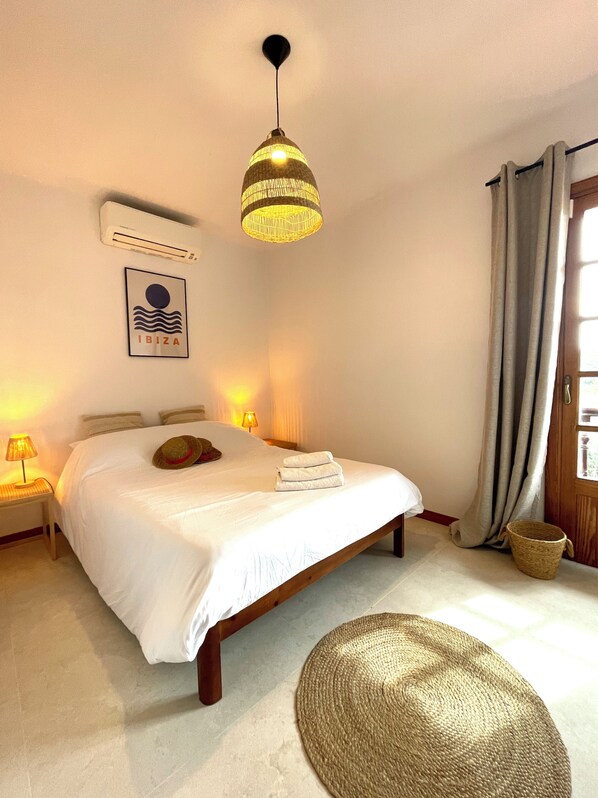 A bedroom with a bed, a hanging lamp, a wall-mounted air conditioner, a framed picture, a door with curtains, and a woven basket. - Authentic & spacious Villa with amazing pool (Santa Gertrudis de Fruitera)