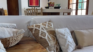 A living room with a white sofa, woven throw pillows, a wooden coffee table, a red abstract painting, and a dining area with a wooden table and chairs.