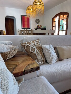Ein Wohnzimmer mit einem weißen Sofa, gewebten Zierkissen, einem Holztisch, einem roten abstrakten Gemälde und einem Essbereich mit Holztisch und Stühlen.