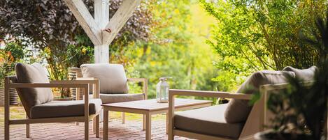Un patio con mesa de madera y dos sillas acolchadas, rodeado de plantas en macetas y un piso de ladrillo.