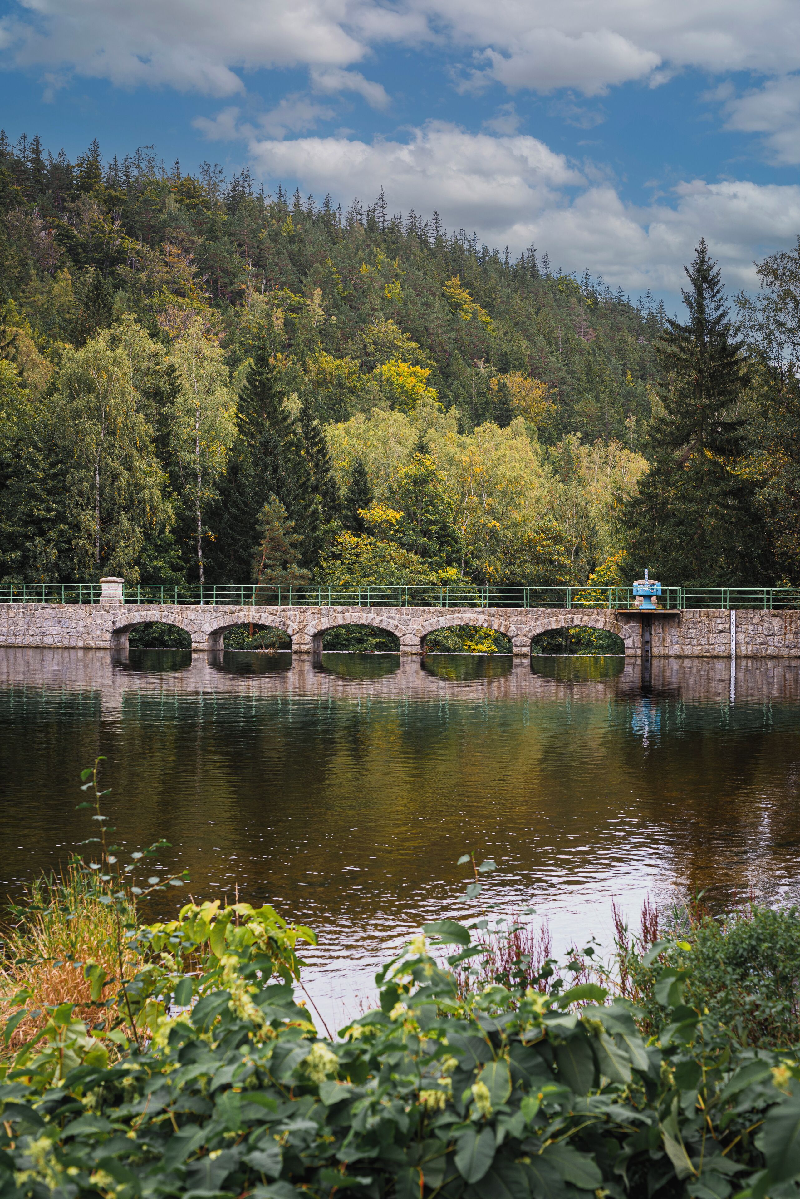 A stone bridge over a calm body of water, surrounded by lush greenery and trees.