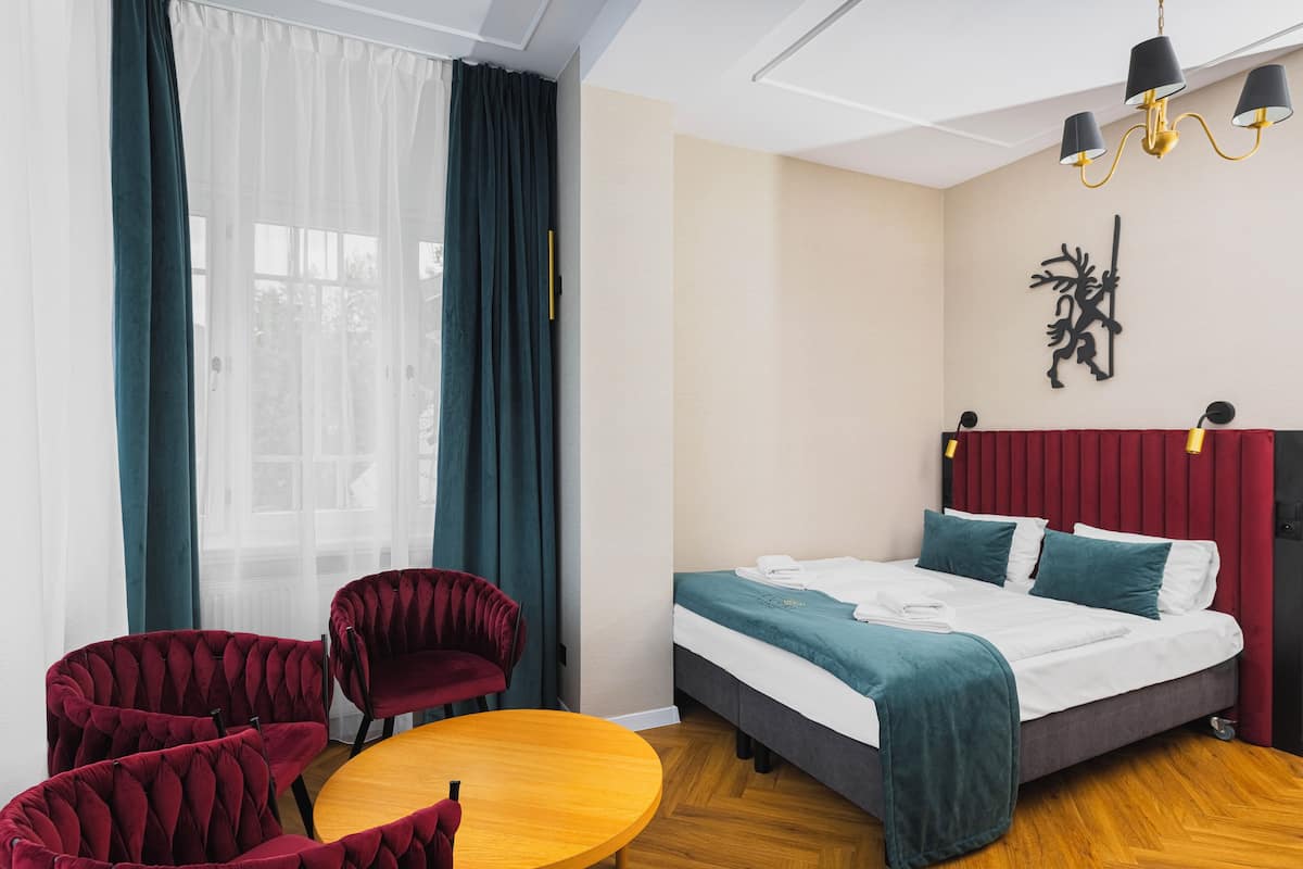 Quarto de hotel com uma cama, duas poltronas de veludo vermelho, uma mesa de madeira e uma janela grande com cortinas leves.