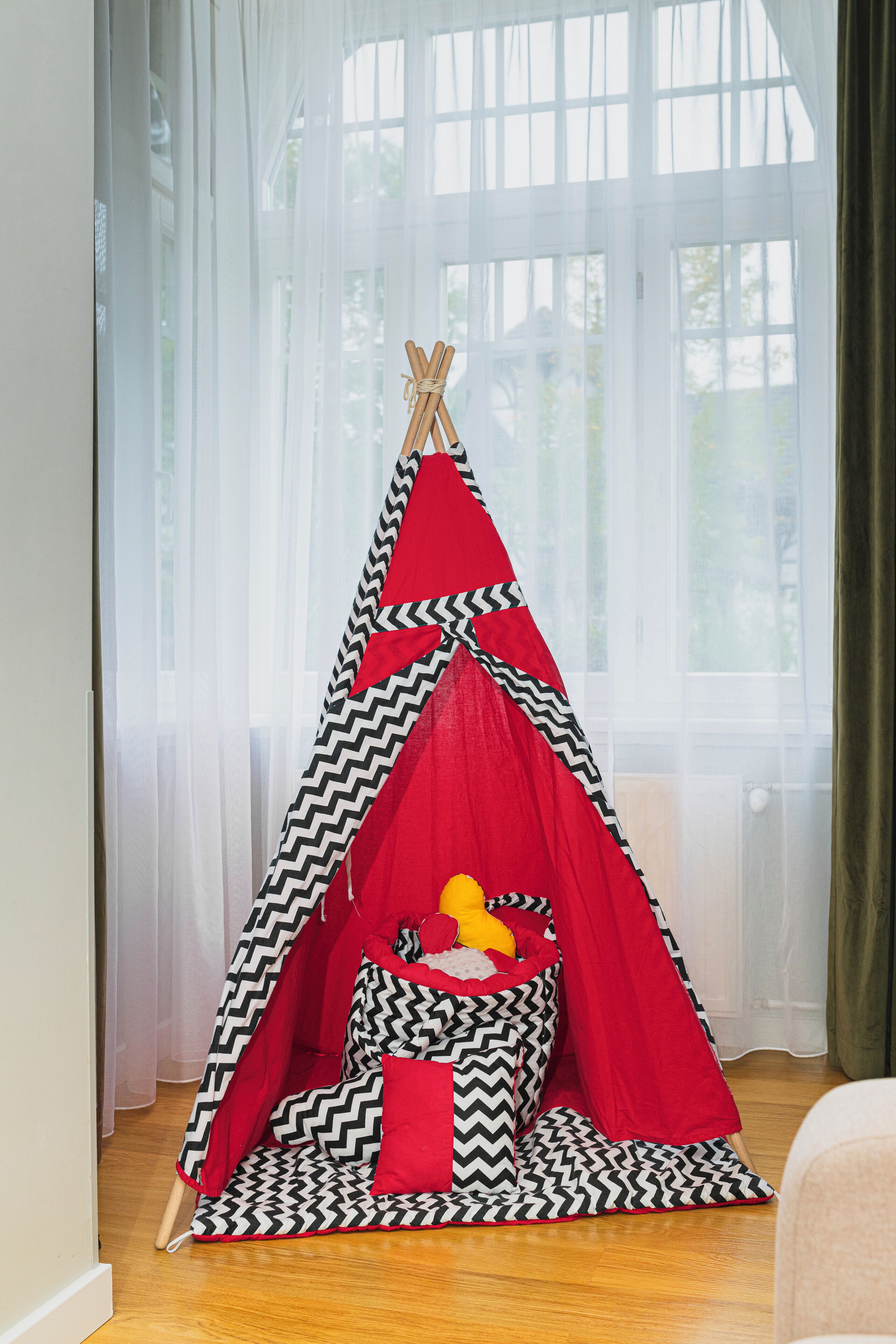 Uma tenda tipi com padrão em vermelho e preto, com uma cama pequena no interior, posicionada em um quarto com uma janela grande e cortinas translúcidas.