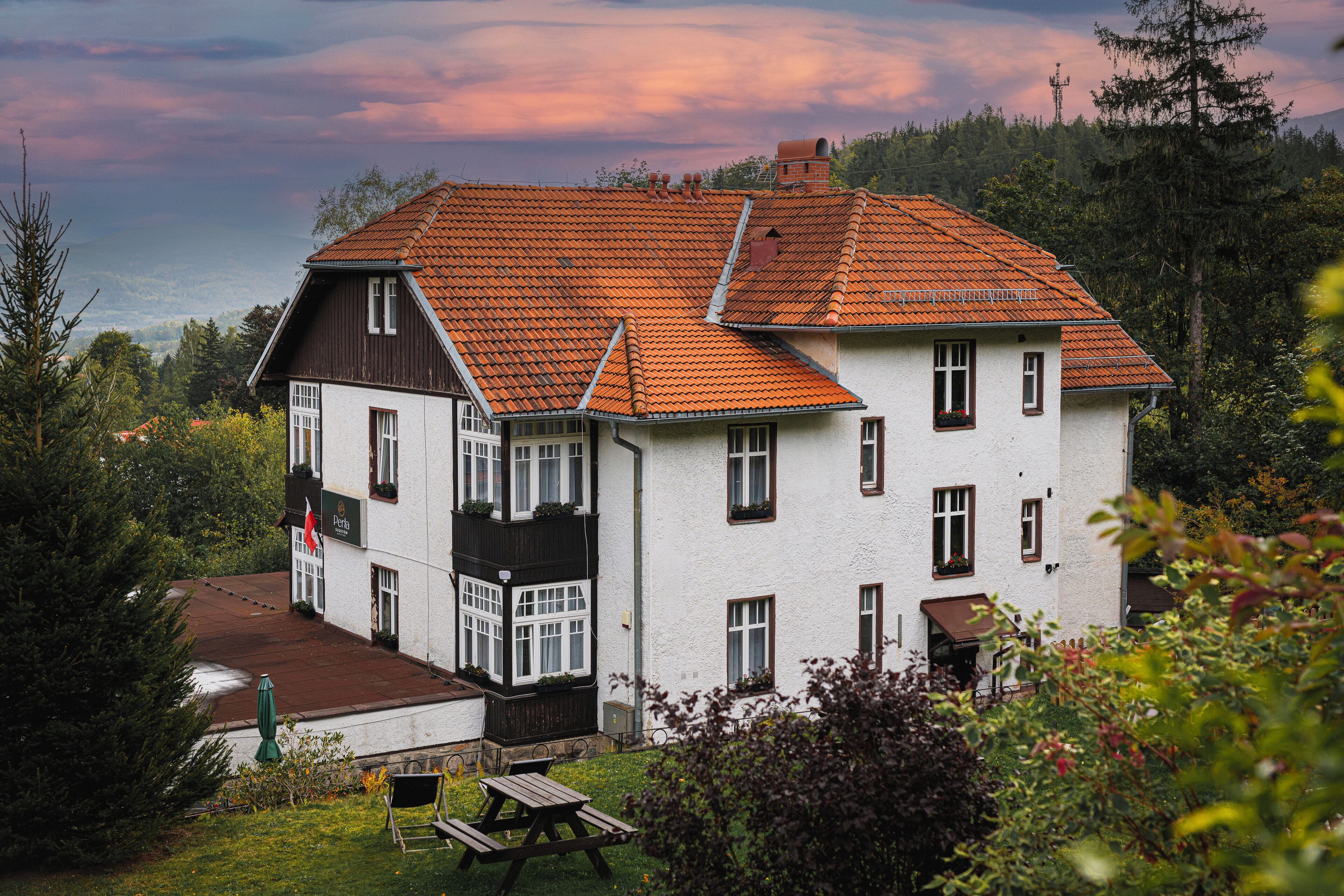 A two-story building with a red-tiled roof and white walls, surrounded by greenery and trees.