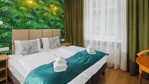 Habitación de hotel con una cama, dos mesitas de noche, una silla de madera, una ventana con cortinas traslúcidas y un papel tapiz con hojas verdes.