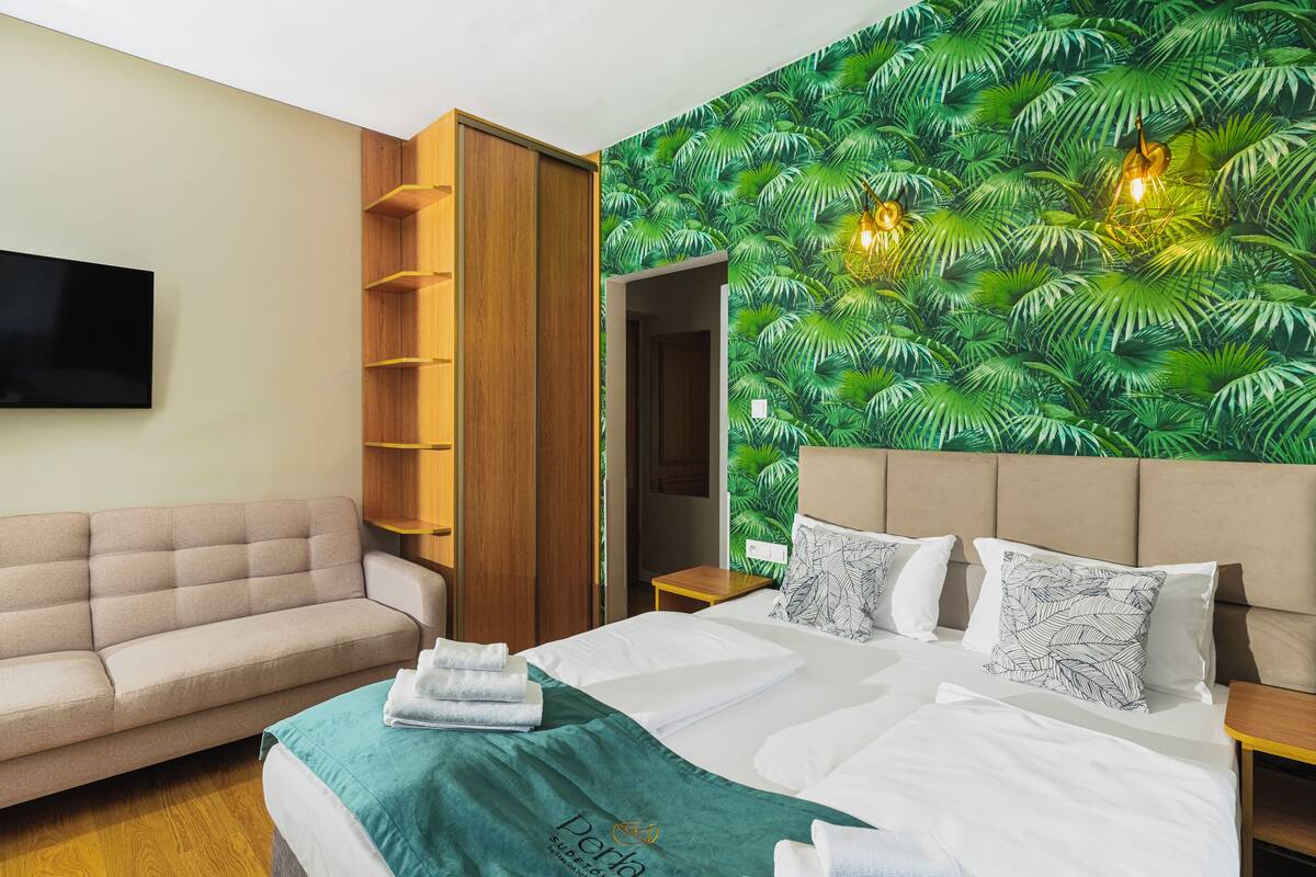 Quarto de hotel com cama, sofá, guarda-roupa de madeira, televisão e parede com padrão de folhagem.