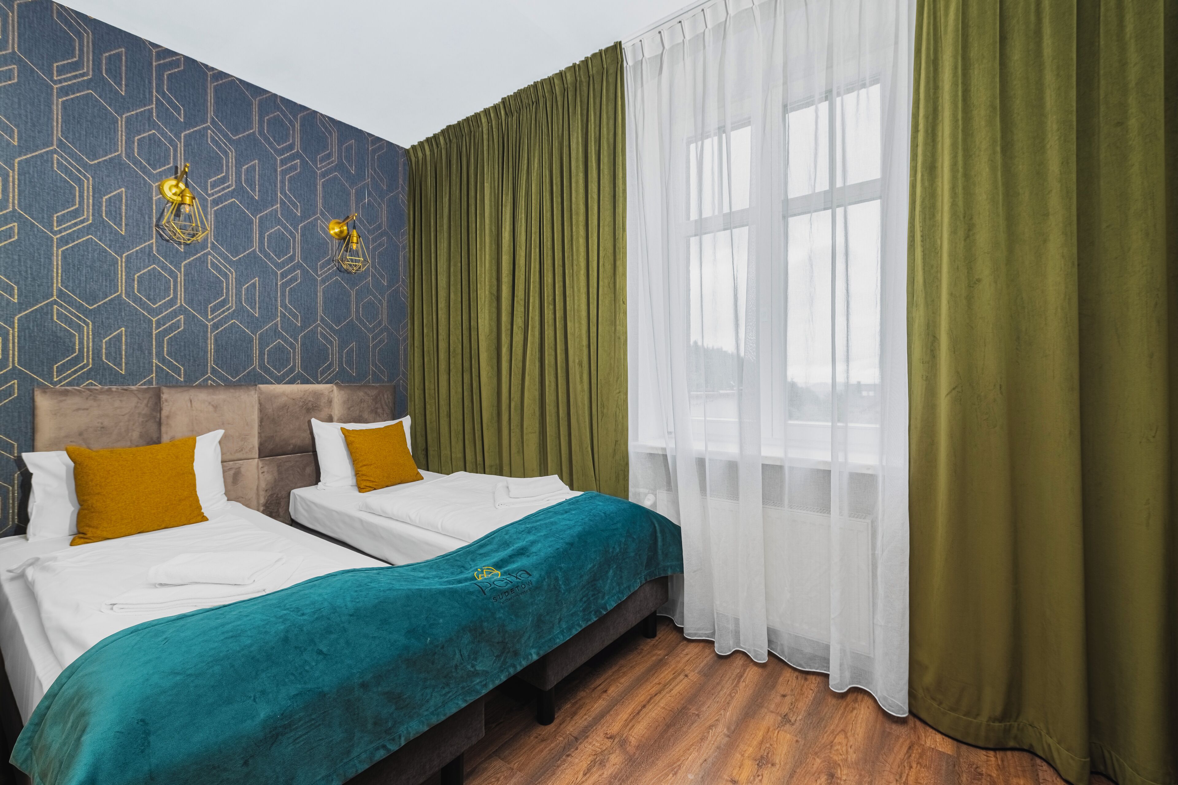 Une chambre d’hôtel avec deux lits, un mur décoré d’un motif et une grande fenêtre avec des rideaux légers.