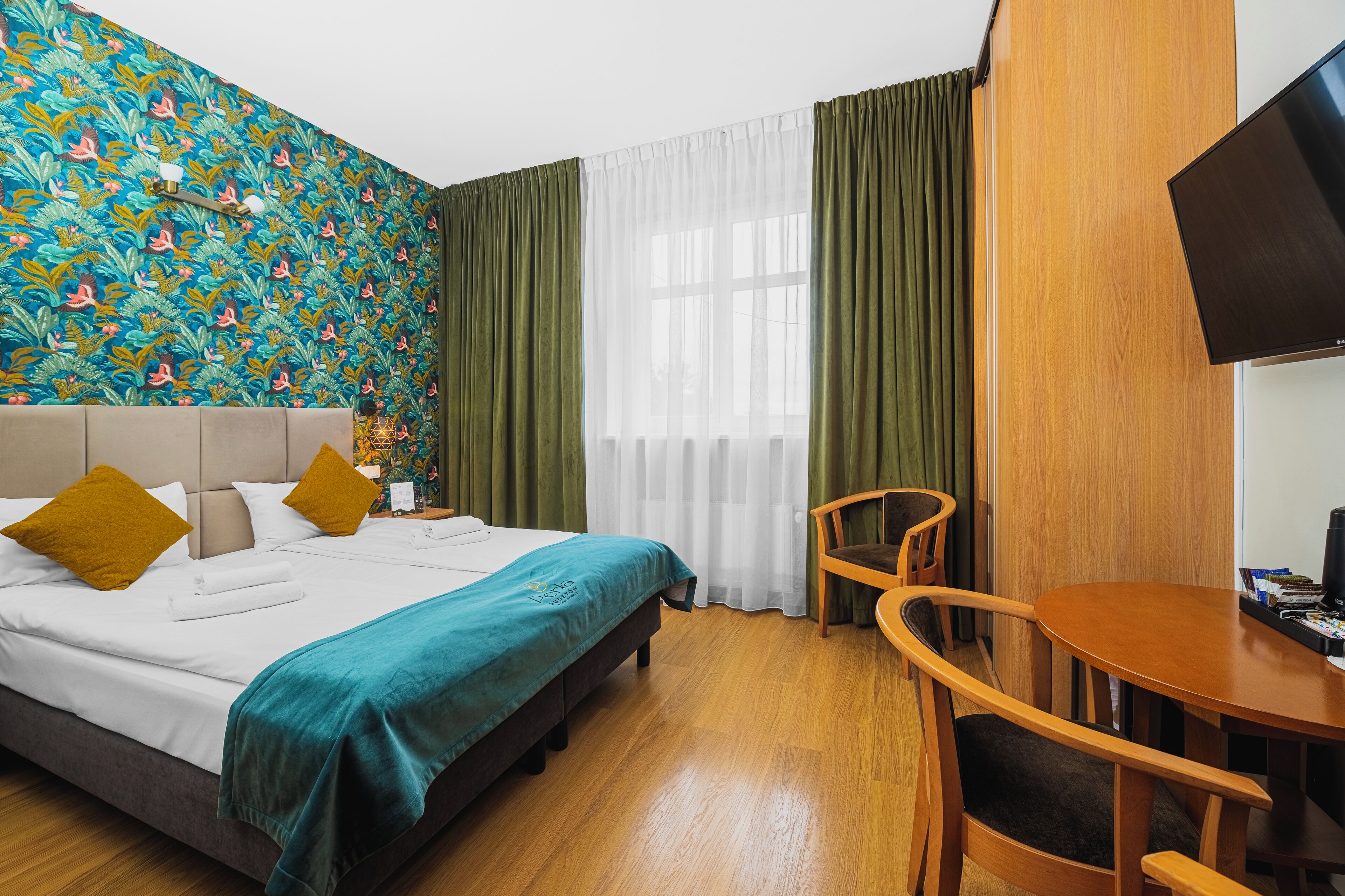 A hotel room with a bed, a wooden chair, a small table, a TV, and a window with curtains.