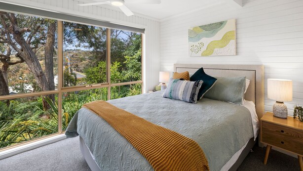 A bedroom with a large window, a bed with a grey and mustard-colored blanket, a ceiling fan, and a wall-mounted lamp. - Soulhaven (Anglesea)