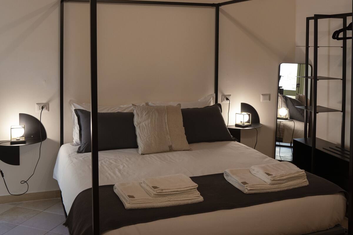 A four-poster bed with white linens, a black metal frame, and a small bedside table with a lamp.