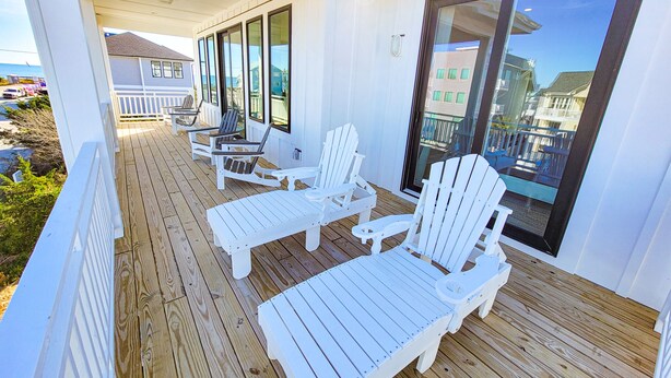 A covered outdoor deck with white Adirondack chairs and a table, overlooking a residential area and the ocean. - Gone Coastal: A Paws-itively Perfect Beach Escape! (Surf City)