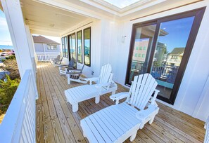 A covered outdoor deck with white Adirondack chairs and a table, overlooking a residential area and the ocean. - Gone Coastal: A Paws-itively Perfect Beach Escape! (Surf City)