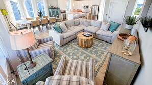 A spacious living area with a large sectional sofa, a coffee table, and a dining area with a table and chairs. There is a kitchen in the background with modern appliances. - Gone Coastal: A Paws-itively Perfect Beach Escape! (Surf City)