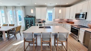 A modern kitchen with white cabinets, a large island with bar stools, stainless steel appliances, and a view of the outside. - Gone Coastal: A Paws-itively Perfect Beach Escape! (Surf City)