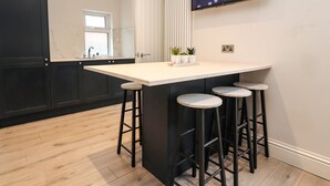 A kitchen with a central island, black bar stools, a white countertop, and a TV mounted on the wall. - 37 Nutter Road (Thornton-Cleveleys)