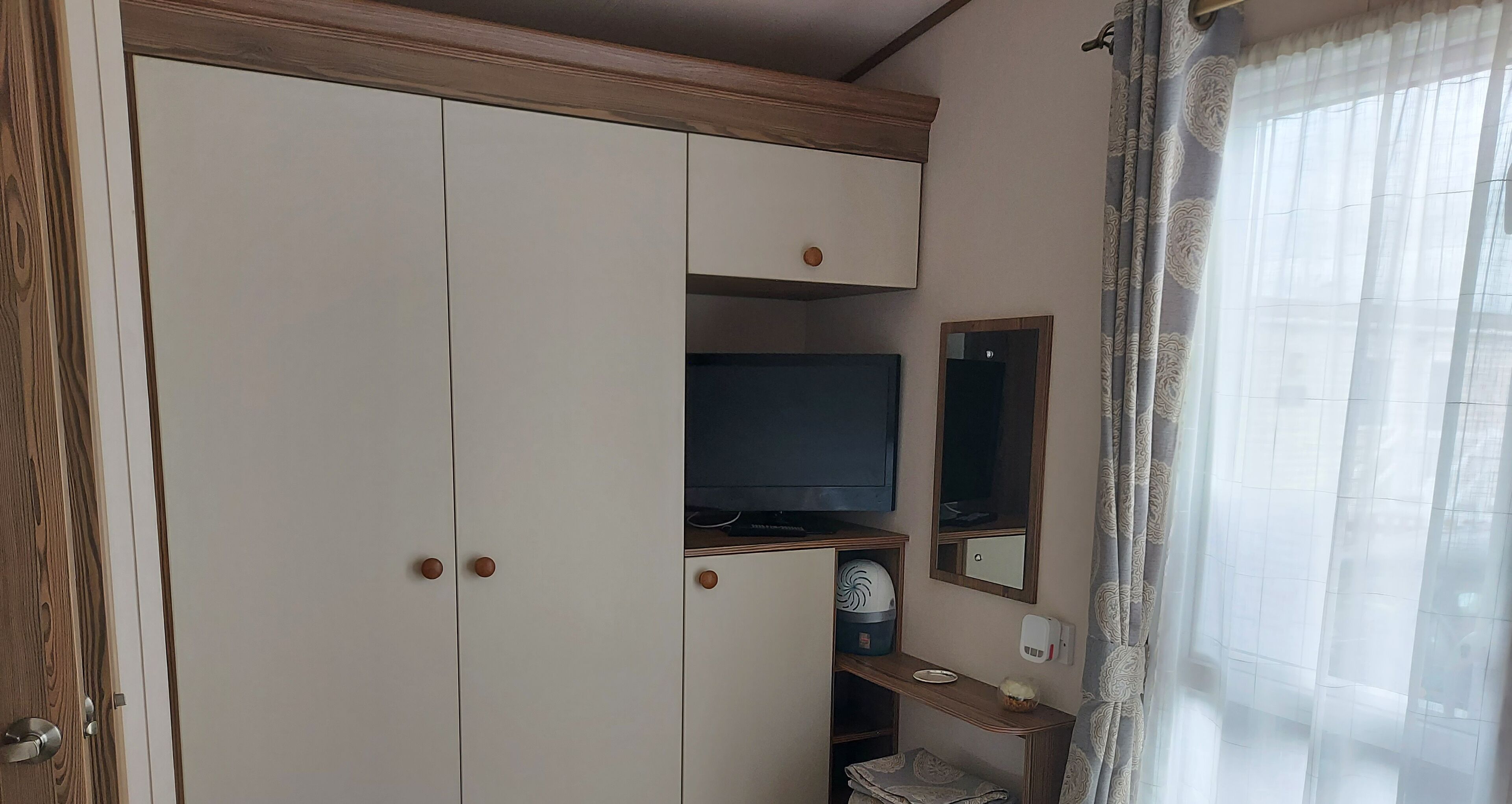 A room with a wardrobe, a television, a mirror, a small table with a bowl, and a window with curtains.