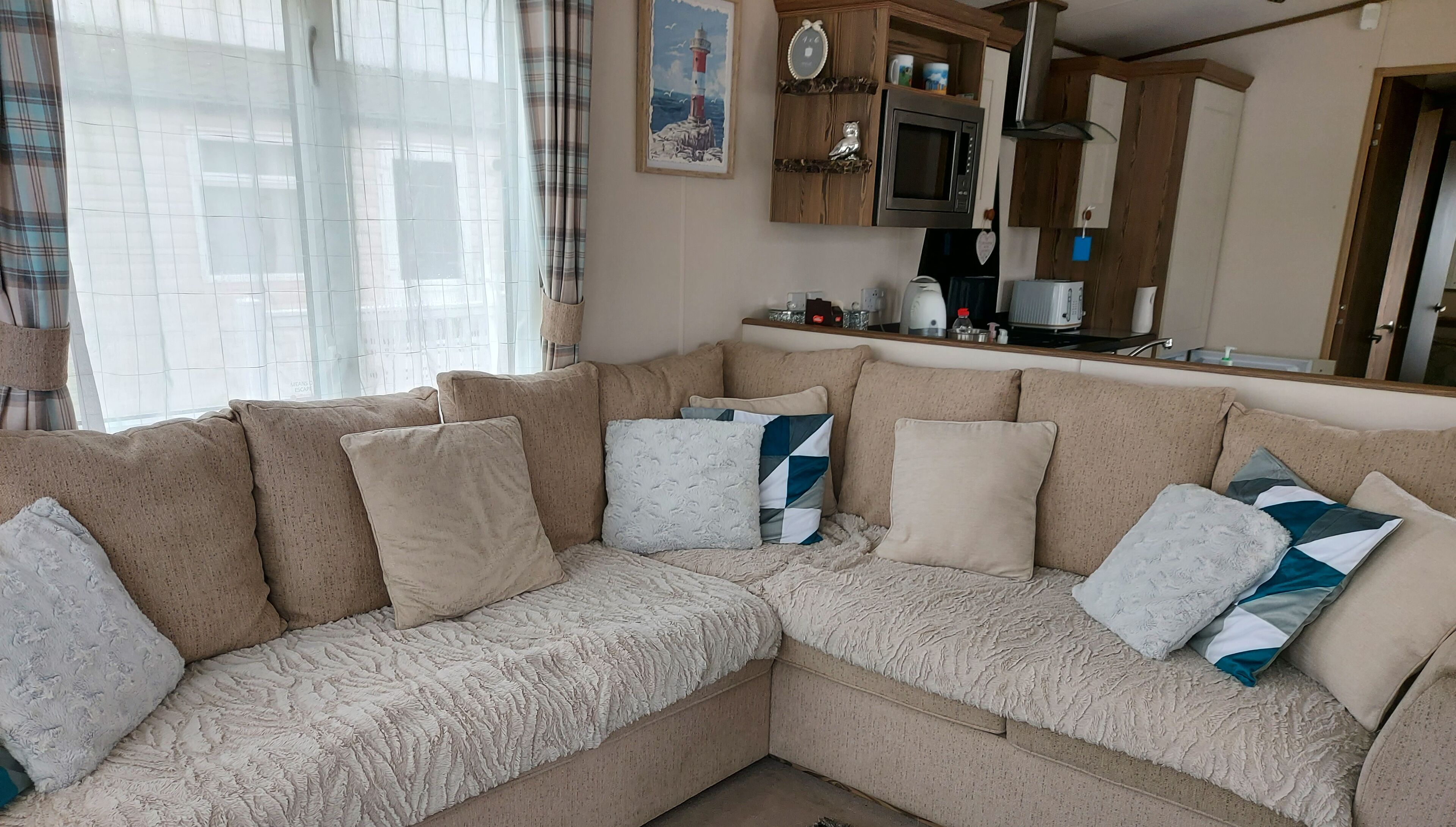 A cozy living area with a beige sectional sofa, a kitchen with wooden cabinets, and a large window with sheer curtains.