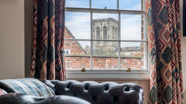A room with a large window, a tufted leather sofa, patterned curtains, and a view of a historic building. - Apartment 4 - The Kings -  a family break that sleeps 4 guests  in 2 bedrooms (York)
