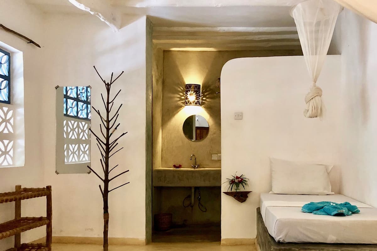 A bedroom with a bed, a bedside table, a window with decorative shutters, a wall-mounted mirror, and a small shelf with a plant.