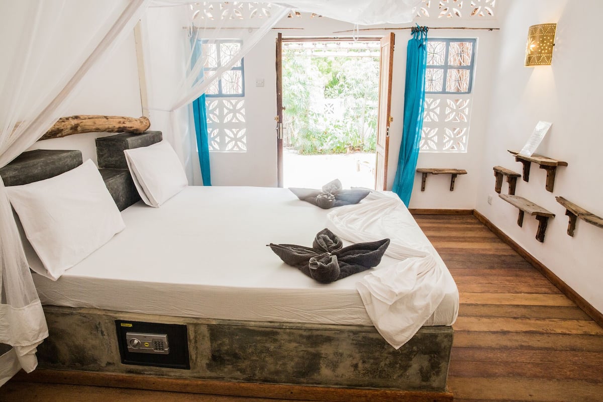 A bed with white and gray bedding, a wooden headboard, and a canopy. There are two small wooden shelves on the wall and a window with blue curtains.