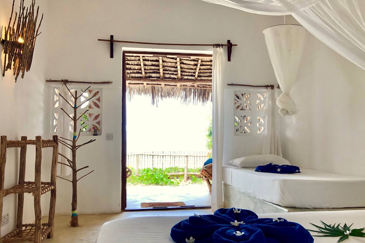 A room with a bed, a wooden chair, a woven door, and a view of greenery outside.