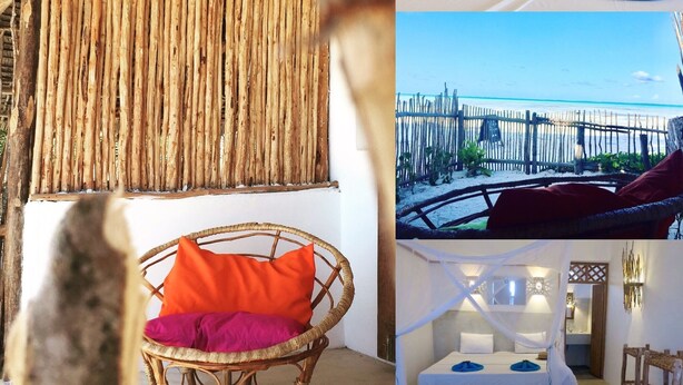 Una habitación con pared de paja, una silla de mimbre con cojín, una cama con dosel y vista a una playa con valla de madera.