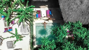 Vista aérea de una zona de piscina con una persona nadando, rodeada de construcciones de techo de paja y abundante vegetación.
