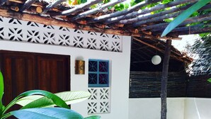 Un edificio pequeño con paredes blancas, una puerta de madera y una ventana con un diseño geométrico. En primer plano, hay un banco de madera y una planta en maceta.