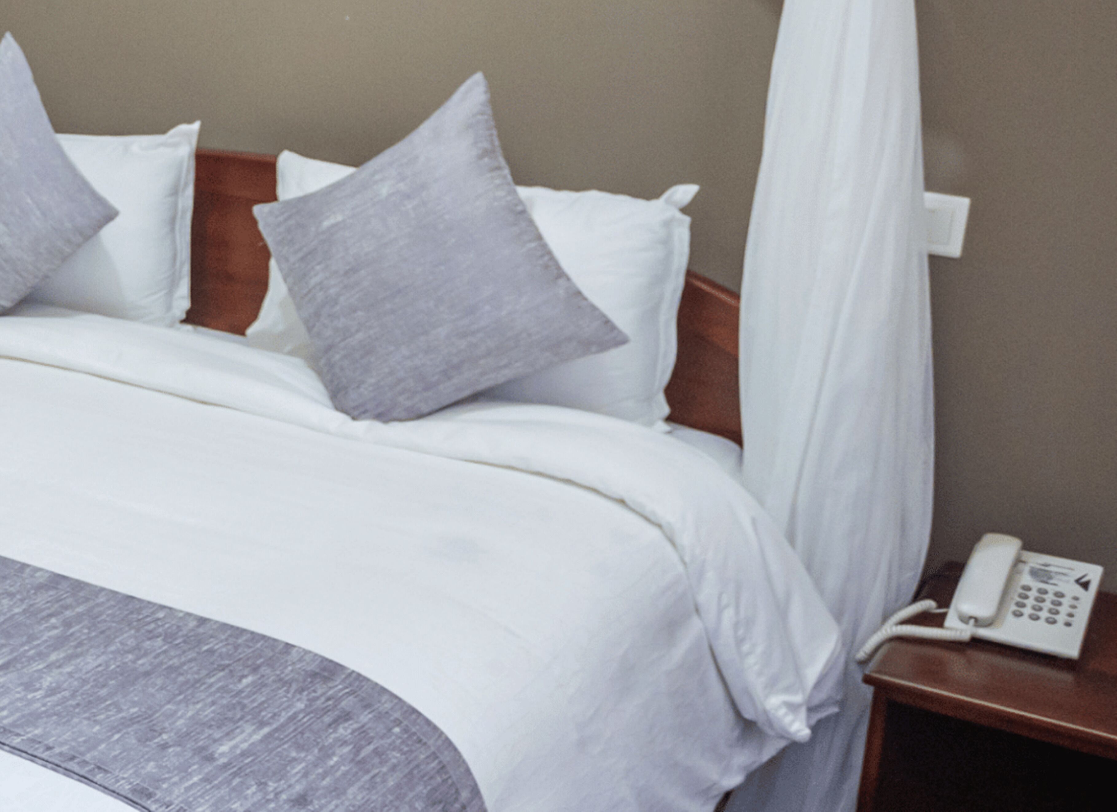 A neatly made bed with white bedding and grey pillows, a wooden headboard, and a bedside telephone on a wooden nightstand.