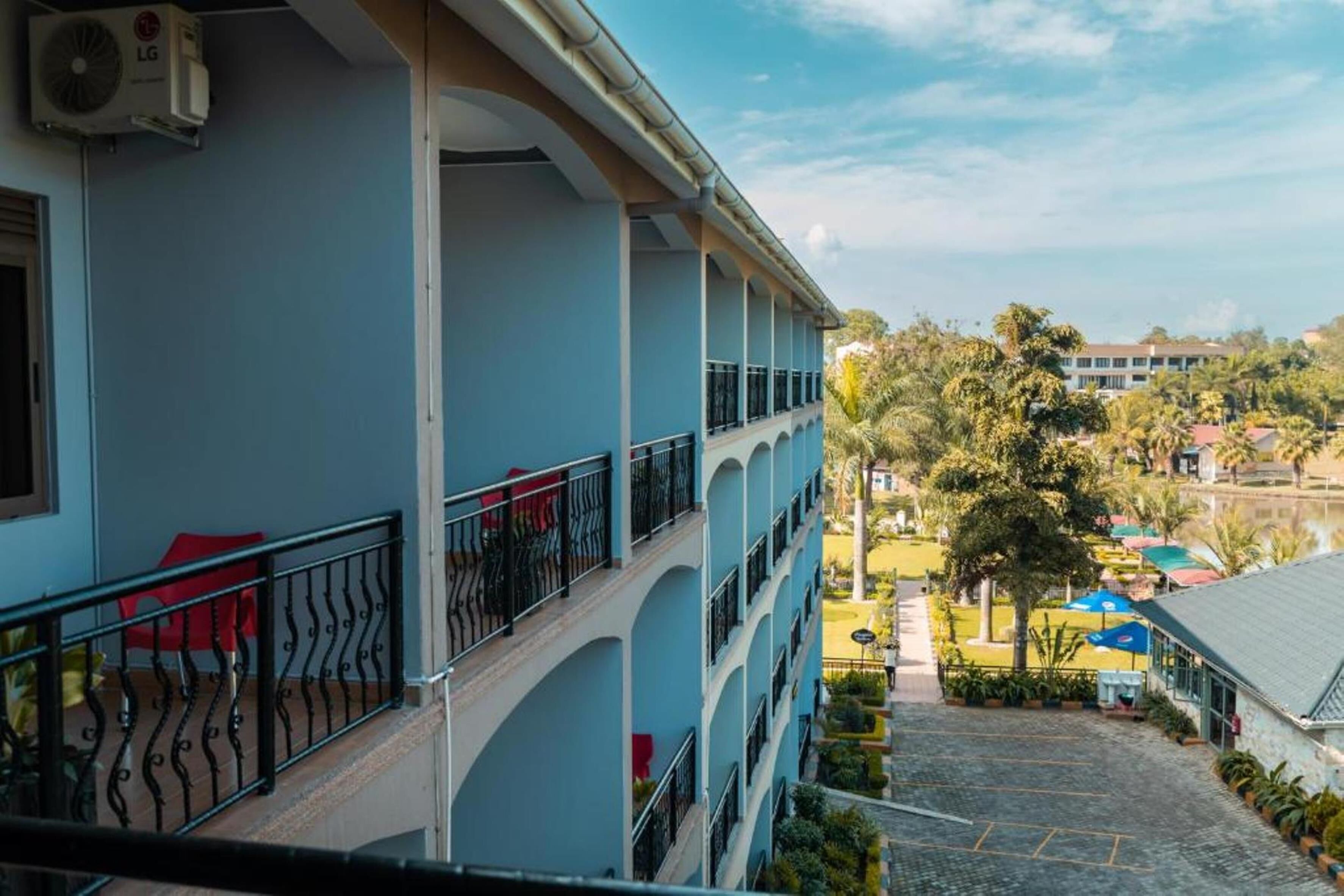 A multi-story hotel with balconies, a view of a landscaped area, and a body of water in the background.