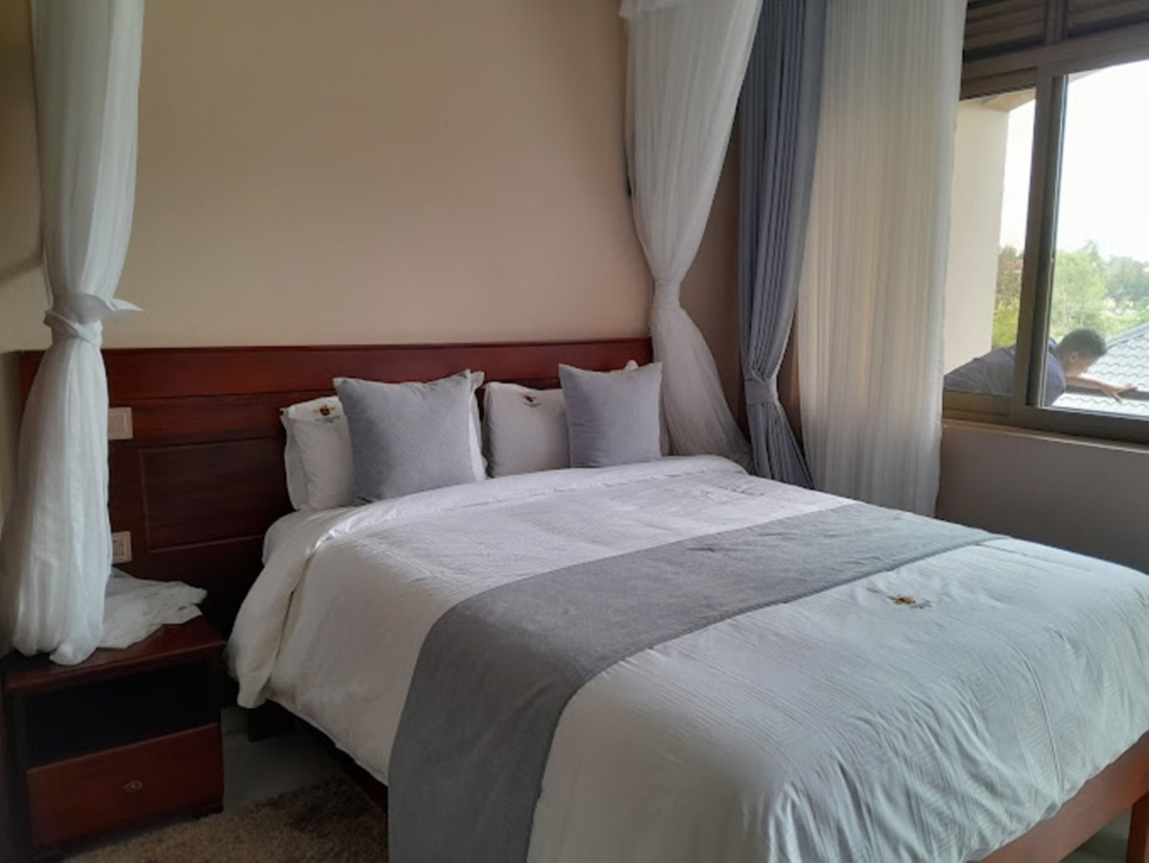 A neatly made bed with white bedding and a grey accent, a wooden headboard, and a window with curtains.