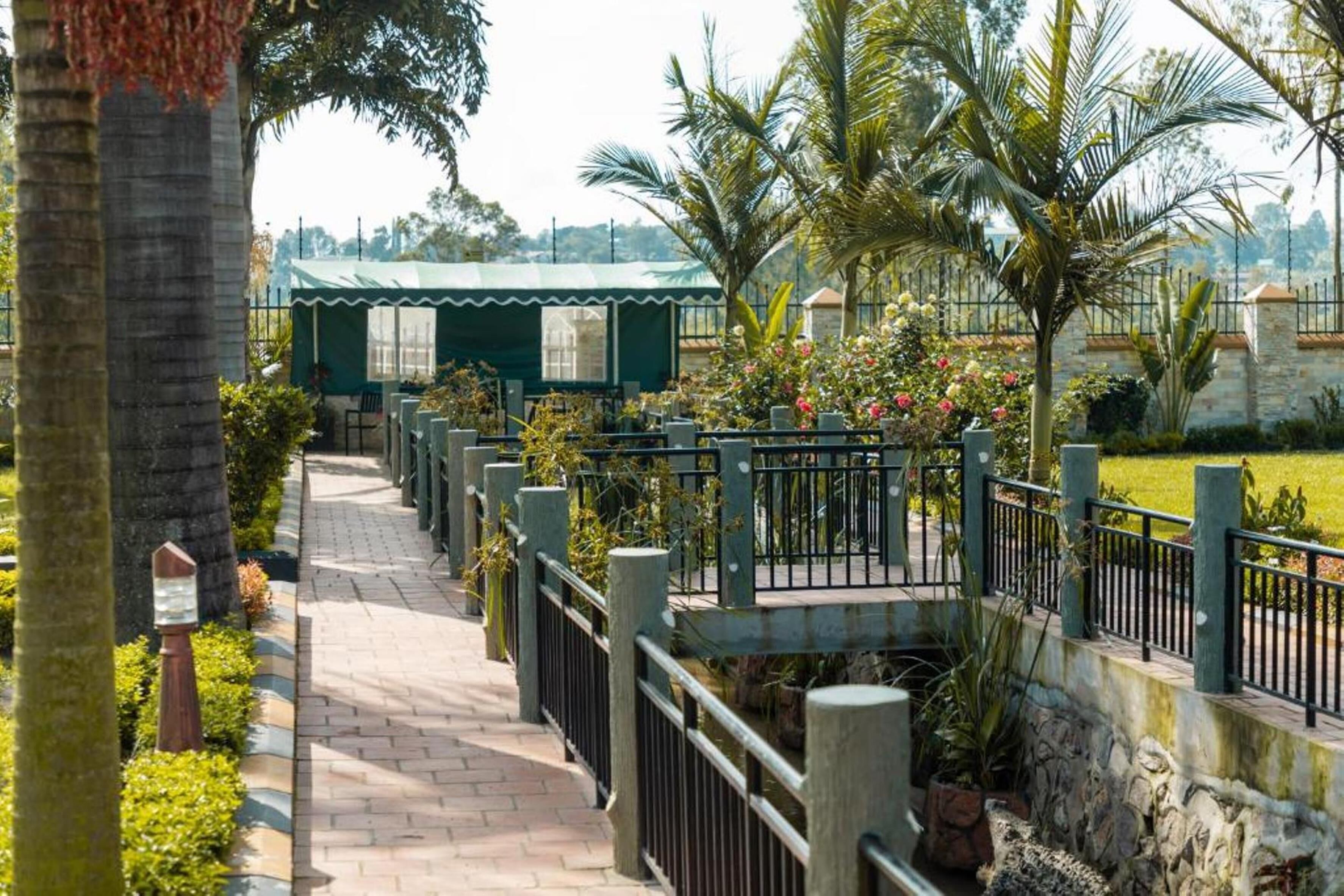 A pathway leading to a green-roofed structure with outdoor seating, surrounded by lush greenery and a stone wall.
