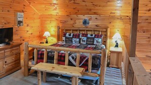 Une pièce chaleureuse, aménagée dans un chalet en bois, avec un lit, deux tables de chevet, un banc, une télévision et un panneau d’accueil.
