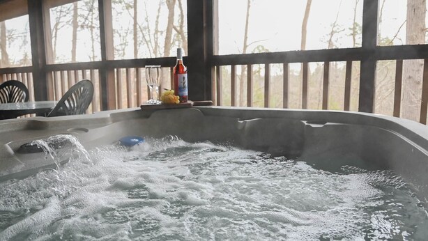Un bain à remous avec de l’eau qui éclabousse, une bouteille de vin et un verre posés sur une table dans un espace extérieur abrité, agrémenté de rampes en bois et de quelques arbres en arrière-plan.