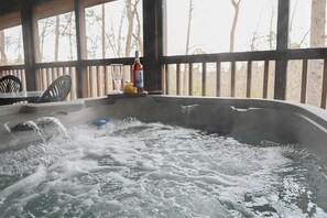 A hot tub with water splashing, a bottle of wine, and a glass on a table in a covered outdoor area with wooden railings and trees in the background. - Cuddly Bear New Hot Tub Private Gas Grill Location (Pigeon Forge)