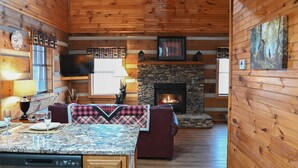 L’intérieur chaleureux d’un chalet avec une cheminée en pierre, des murs en bois, un coin repas doté d’un comptoir en granit et un téléviseur fixé au mur.