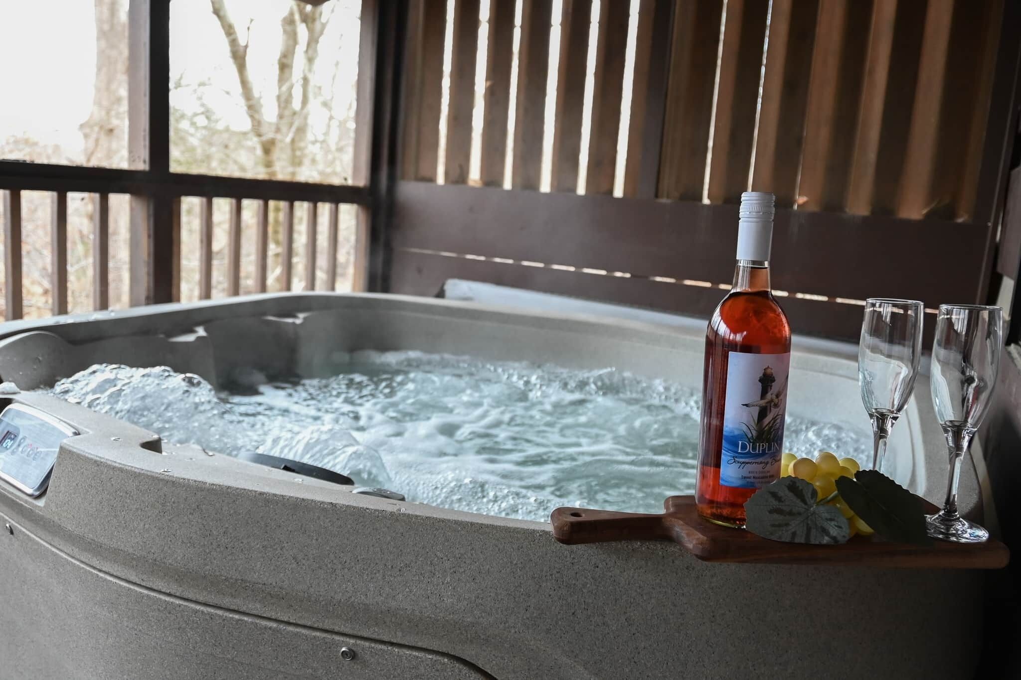 A hot tub with a bottle of Duplin and two glasses, wooden tray, and a wooden railing in the background.