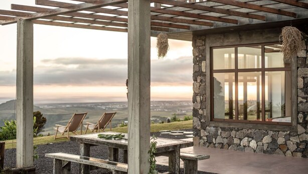 A covered outdoor seating area with a stone wall and large windows, overlooking a scenic landscape. - Villa in Nature (Vila do Porto)