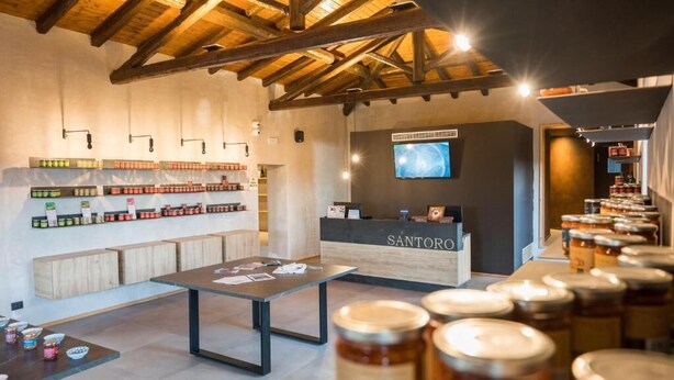 A modern interior with wooden beams, a reception desk labeled 'SANTORO', and shelves of jars.