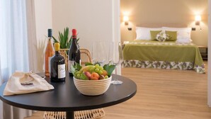 A room with a bed, a table with a bowl of fruit and wine bottles, and a small plant.