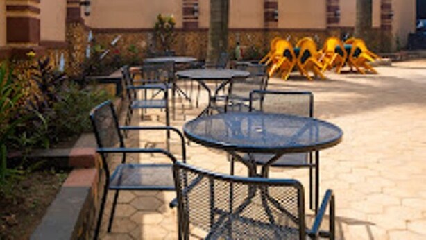 Outdoor seating with tables and chairs, surrounded by a garden and a building with a beige facade. - Governors Hotel (Mukono)