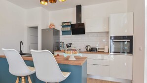 Una cuina moderna amb armaris blancs, un taulell de fusta i un forn integrat. Hi ha dues cadires blanques amb potes de fusta, una illa blava i un plat per a pastissos de vidre.