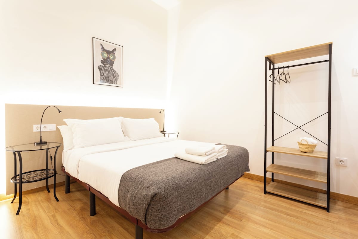 A neatly made bed with a grey blanket, a bedside table with a lamp, a wall-mounted shelf with a basket, and a framed picture of a cat.
