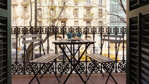 Een balkon met een smeedijzeren hekwerk, twee houten bankjes en een tafel met een bord en een kop.